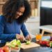 Black woman standing at the counter cutting a cucumber with other vegetables laid out on the counter top, practicing healthy living